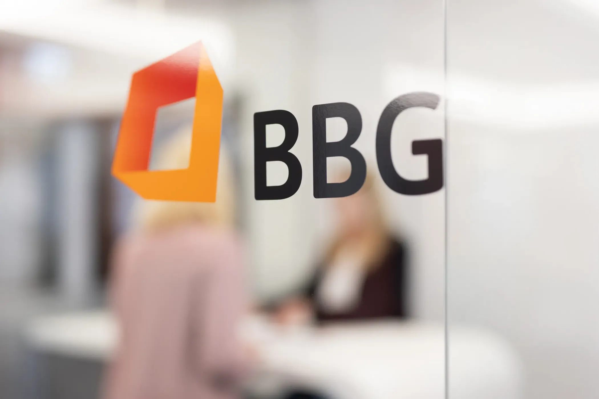 A glass wall with the BBG logo and two indistinct people talking in an office in the background.