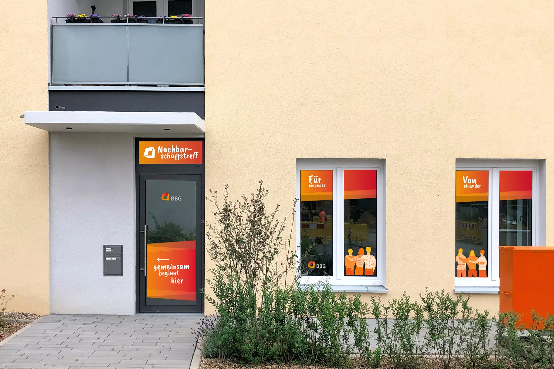 Der Eingang zu einem Gemeindezentrum mit großen Fenstern, Schildern in Deutsch, Orange und Gelb und kleinen Pflanzen im Vordergrund.