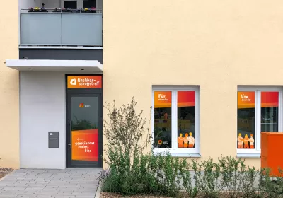 Der Eingang zu einem Gemeindezentrum mit großen Fenstern, Schildern in Deutsch, Orange und Gelb und kleinen Pflanzen im Vordergrund.