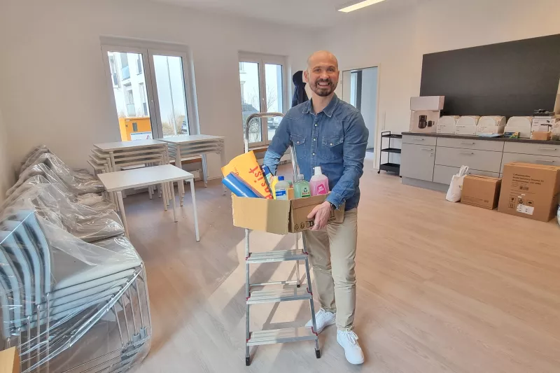 Drinnen steht ein lächelnder Mann mit einer Kiste voller Reinigungsmittel auf einer kleinen Treppe in einem hellen, modernen Raum mit gestapelten Stühlen.