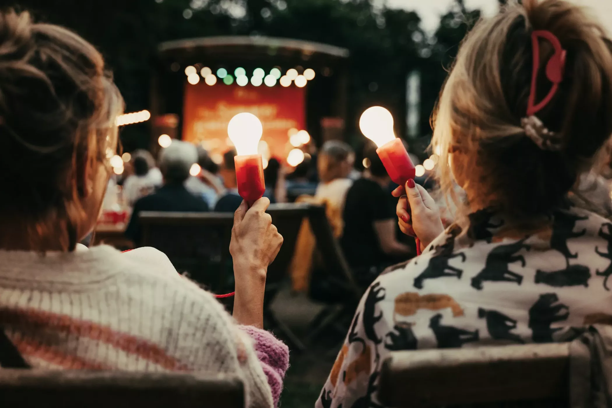Zwei Personen mit Taschenlampen an Stöcken, sitzend bei einem Konzert oder einer Veranstaltung im Freien mit einer Bühne im Hintergrund.