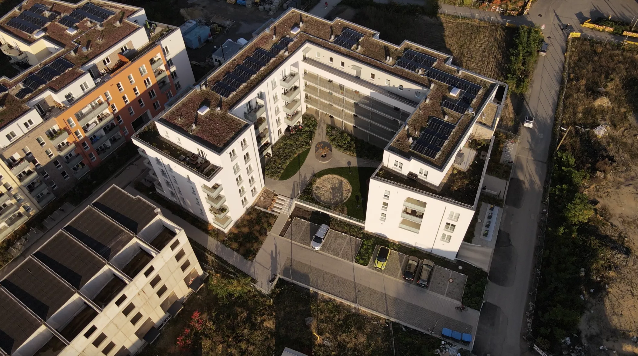 Çatılarında güneş panelleri ve yeşil alanlar ile park edilmiş arabaların bulunduğu merkezi bir avlusu olan modern bir konut kompleksinin havadan görünümü.