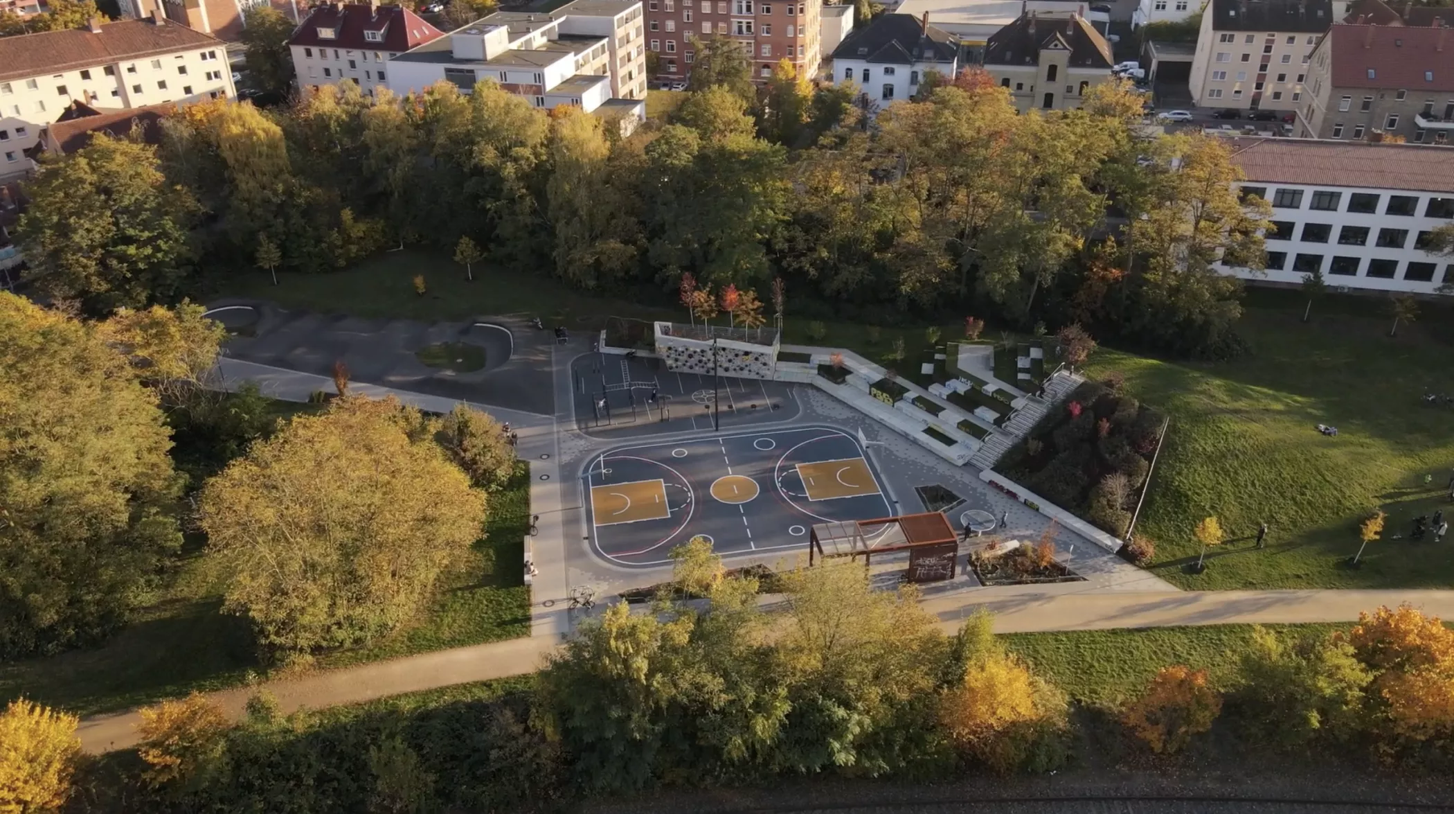 Bir sonbahar gününde ağaçlar ve yakındaki binalarla çevrili bir basketbol sahasının havadan görünümü.