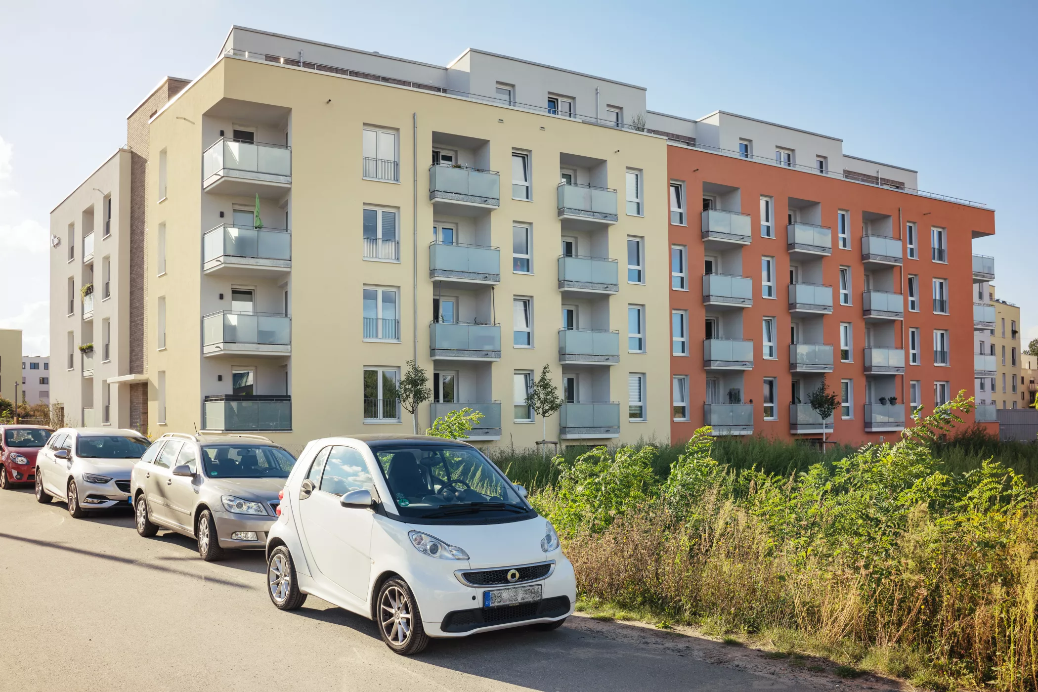 Eine Reihe von Autos, die an einem sonnigen Tag vor modernen Wohnhäusern mit Balkonen auf der Straße geparkt sind.