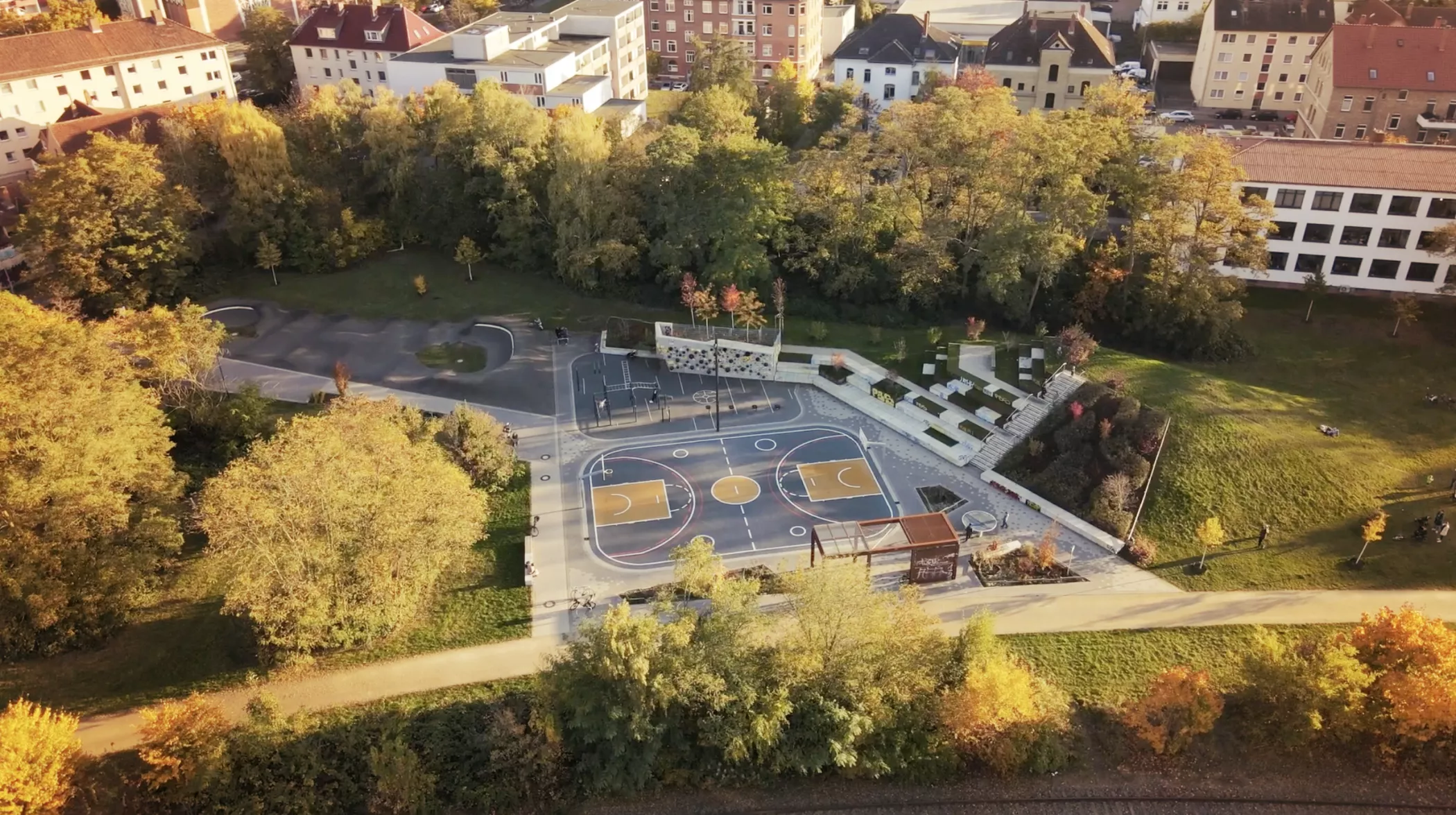 An aerial view of an outdoor basketball court surrounded by trees and buildings on a sunny fall day.