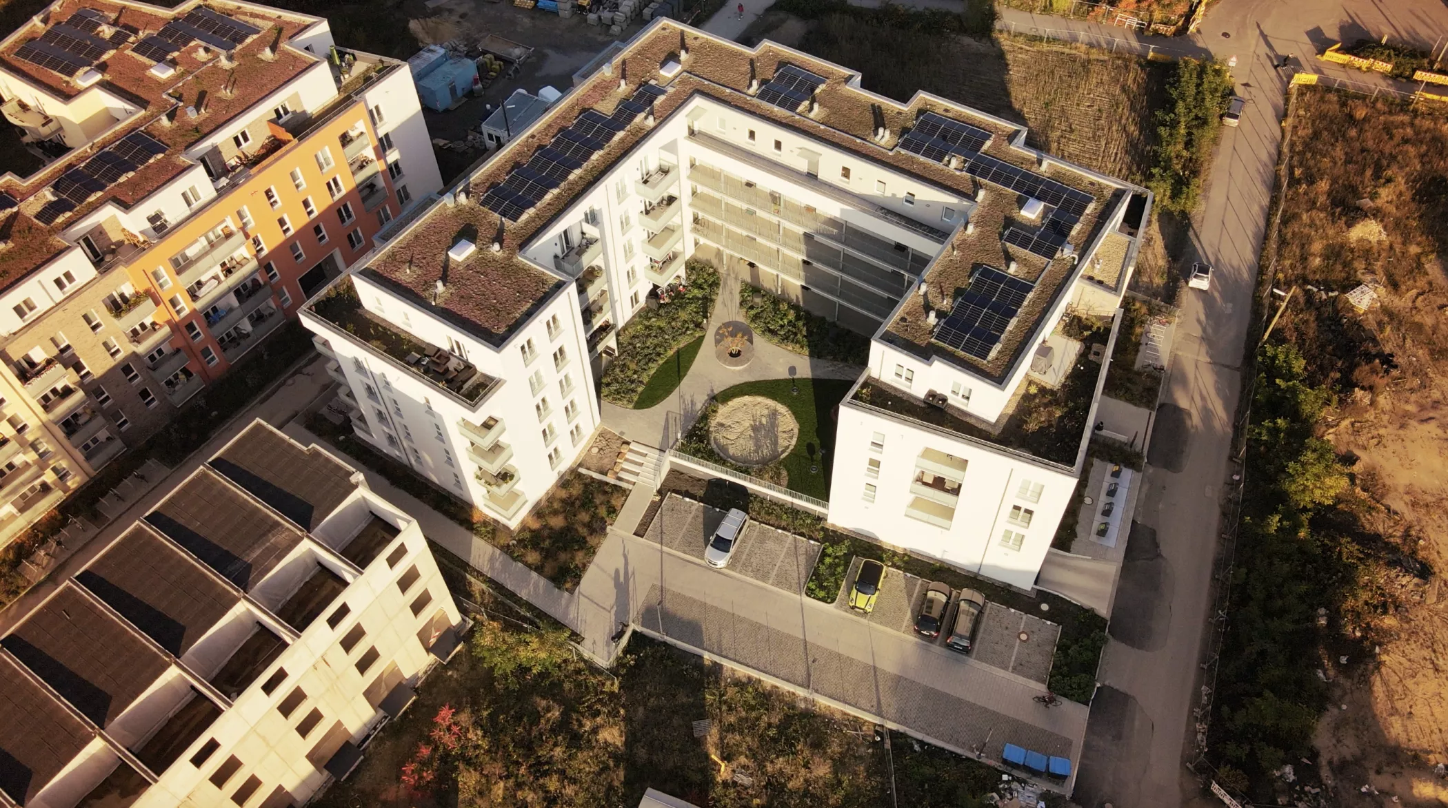 Luftaufnahme eines modernen Wohnkomplexes mit zentralem Innenhof, parkenden Autos und Solarzellen auf dem Dach.