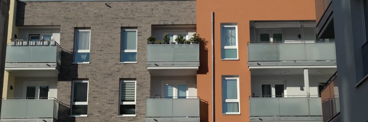 A modern residential building with a gray and orange brick façade, glass balconies and large windows under a clear blue sky.