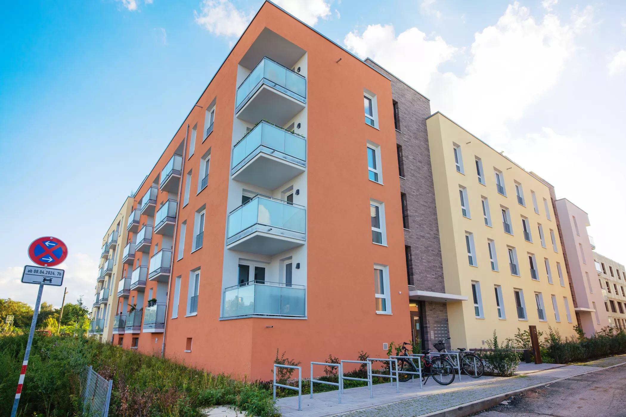 A modern residential building with an orange and yellow façade, glass balconies and bicycles parked near the entrance on a sunny day.