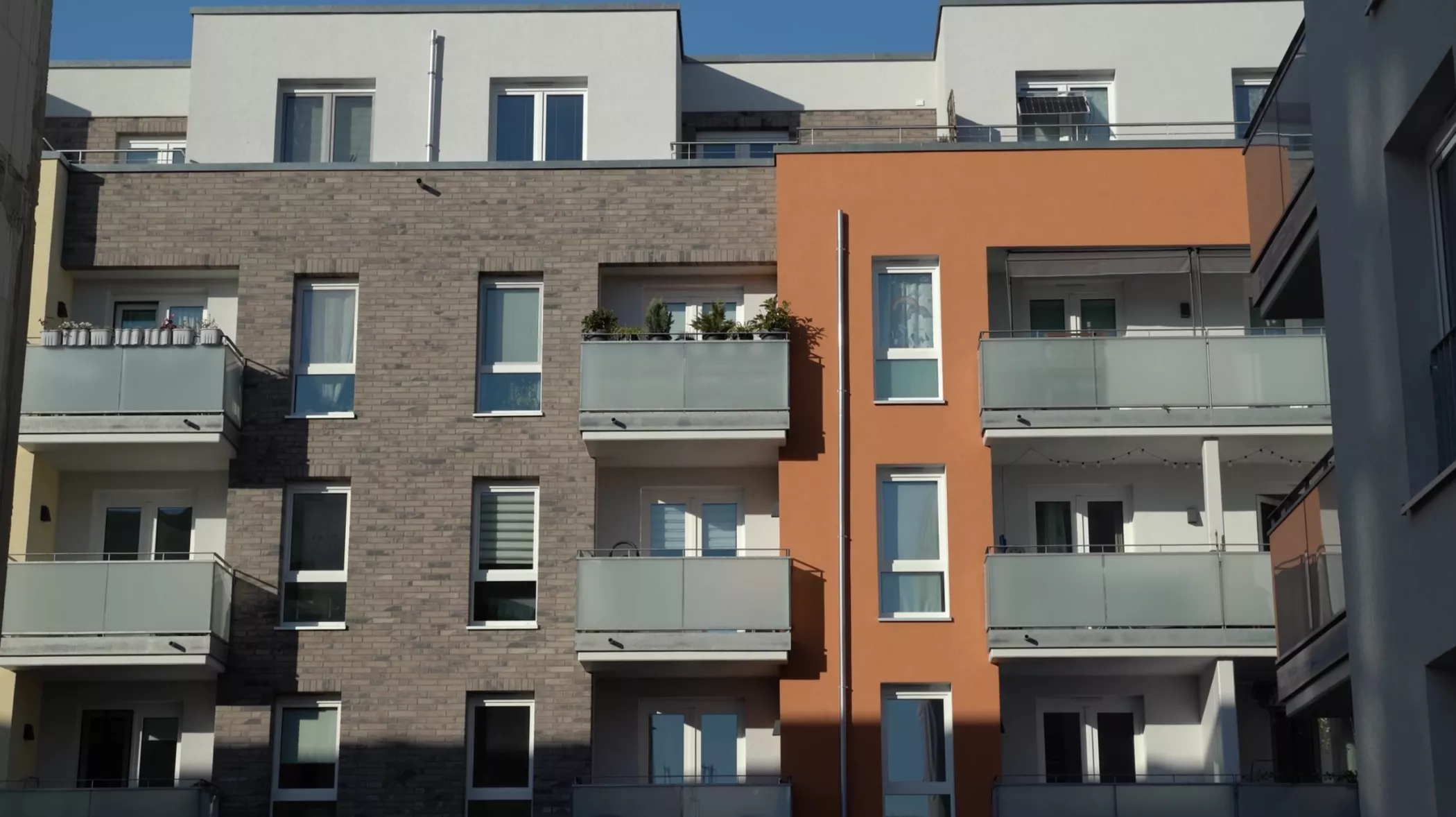 A modern residential building with a gray and orange brick façade, glass balconies and large windows under a clear blue sky.