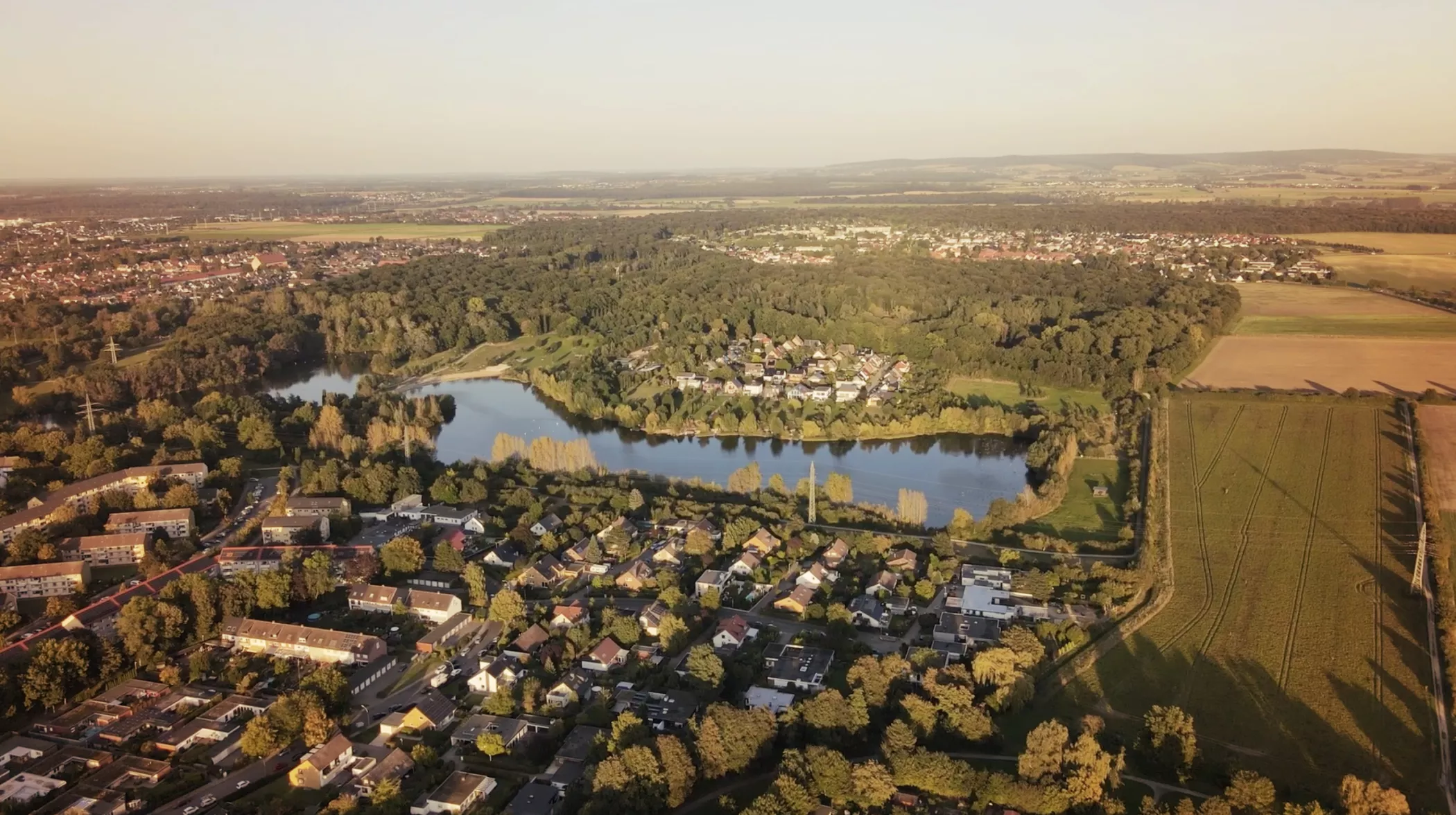 Eine Luftaufnahme einer Stadt mit Häusern, einem See, dichten Bäumen und offenen Feldern unter einem klaren Himmel bei Sonnenuntergang.