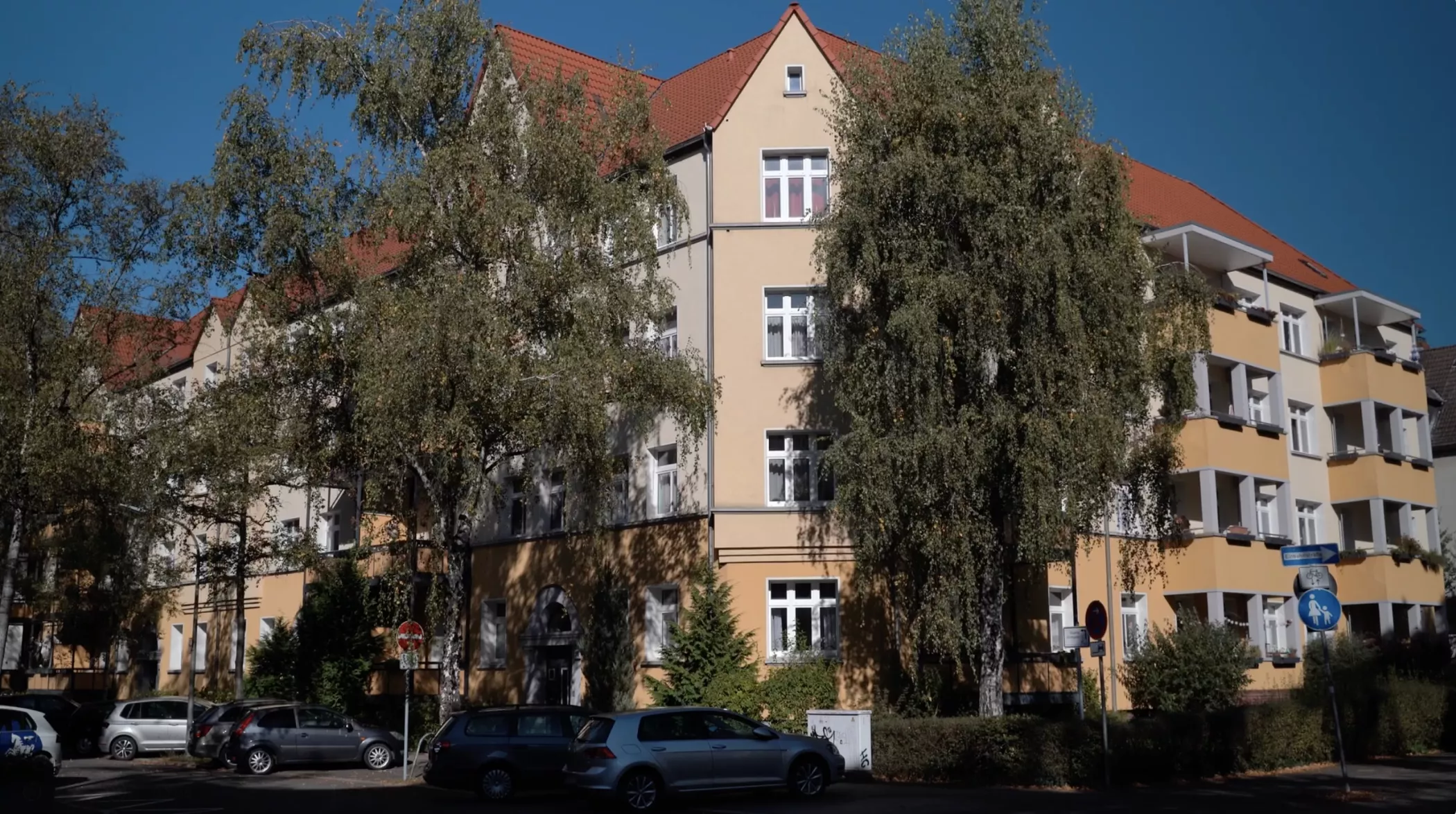 A beige-colored residential building with a red roof, partially concealed by tall trees and with cars parked along the street in front of it.