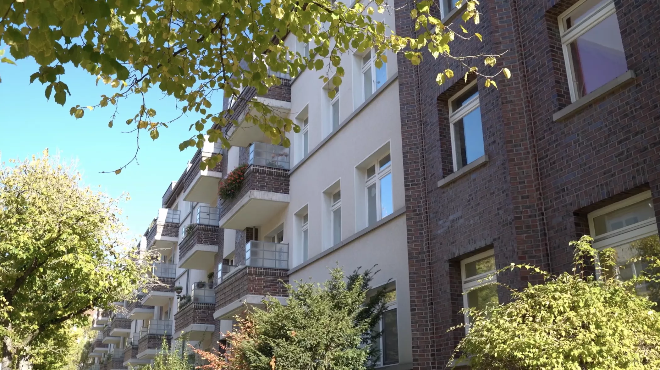 Un immeuble résidentiel de quatre étages avec des balcons entourés d'arbres luxuriants par une journée ensoleillée avec un ciel bleu.