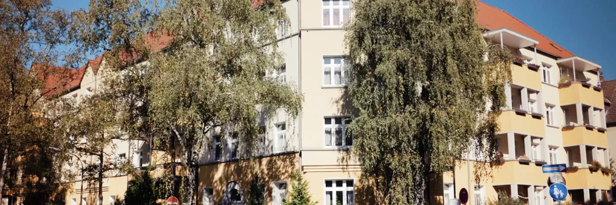 Ein Wohnhaus mit rotem Dach, einer hell erleuchteten Fassade, Bäumen davor und parkenden Autos entlang der Straße an einem sonnigen Tag.