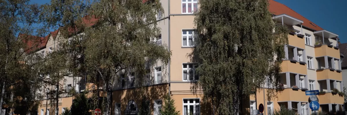 Kırmızı çatılı bej renkli bir konut binası, uzun ağaçlar tarafından kısmen gizlenmiş ve önünde cadde boyunca park etmiş arabalar var.