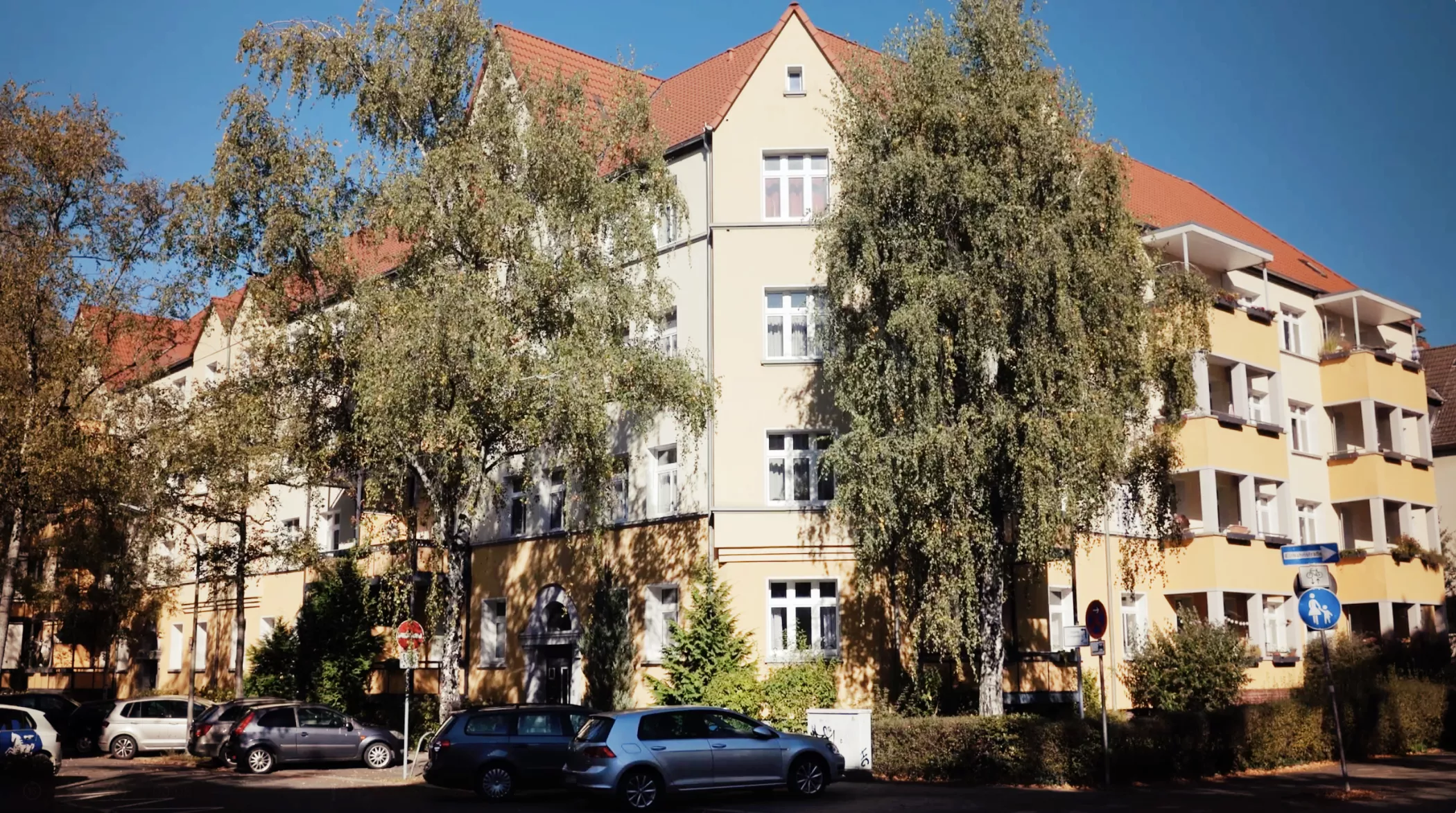 Ein beigefarbenes Wohnhaus mit roten Dächern, davor hohe Bäume und an einem sonnigen Tag parkende Autos entlang der Straße.