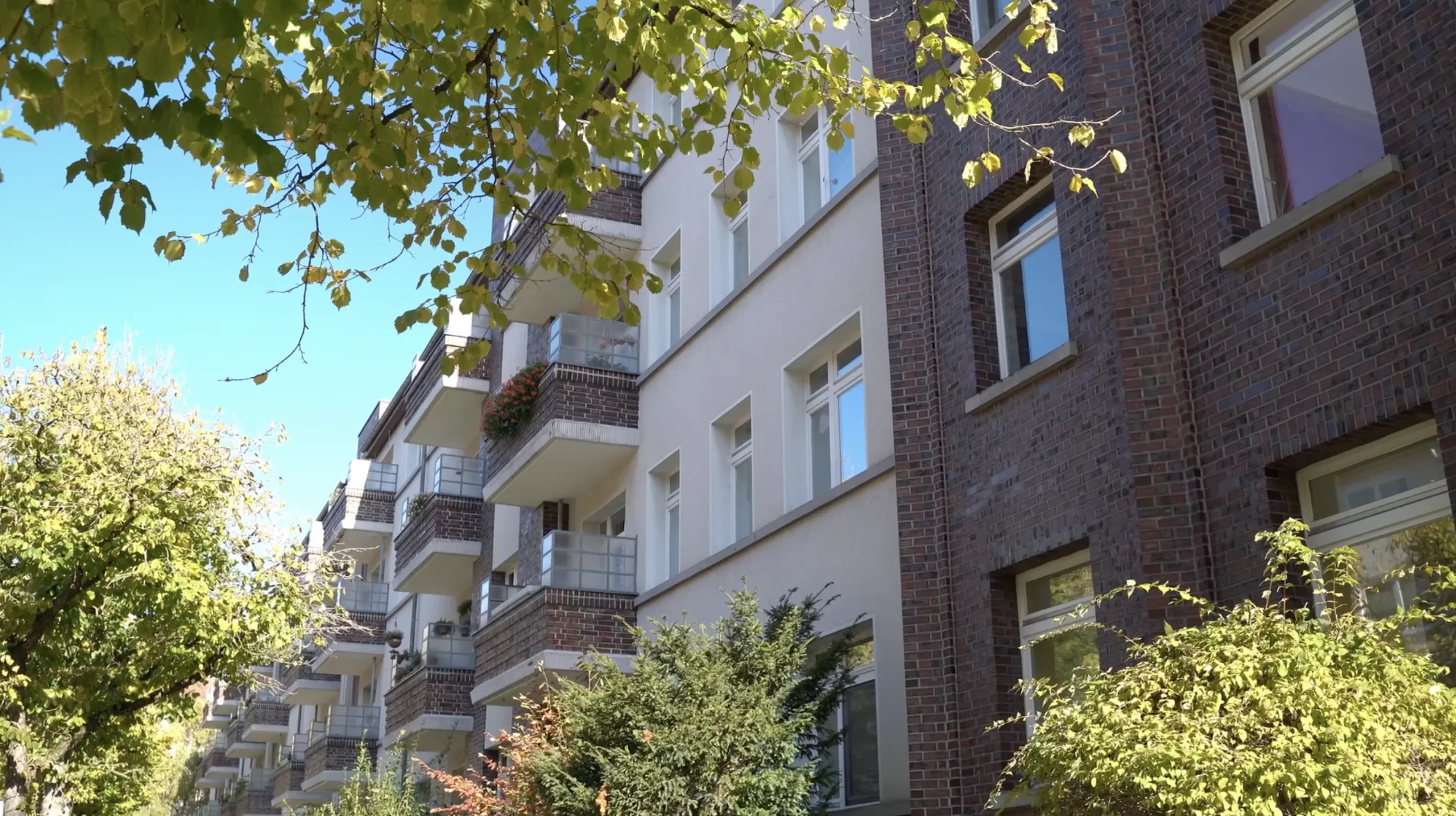 Un immeuble résidentiel de quatre étages avec des balcons entourés d'arbres luxuriants par une journée ensoleillée avec un ciel bleu.