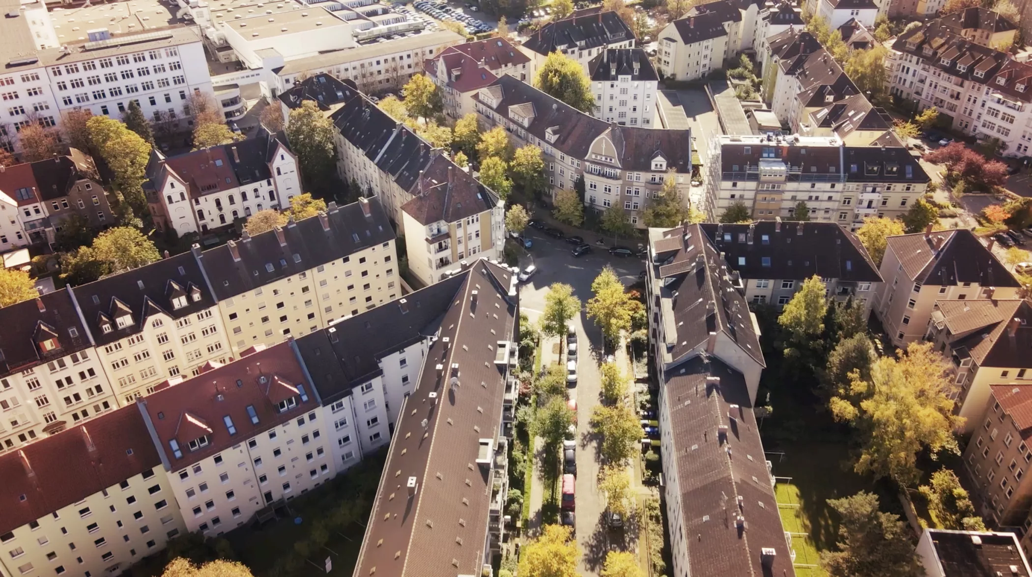 Luftaufnahme eines Wohnblocks in einer europäischen Stadt mit Wohngebäuden, Bäumen und parkenden Autos entlang der Straßen im herbstlichen Sonnenlicht.