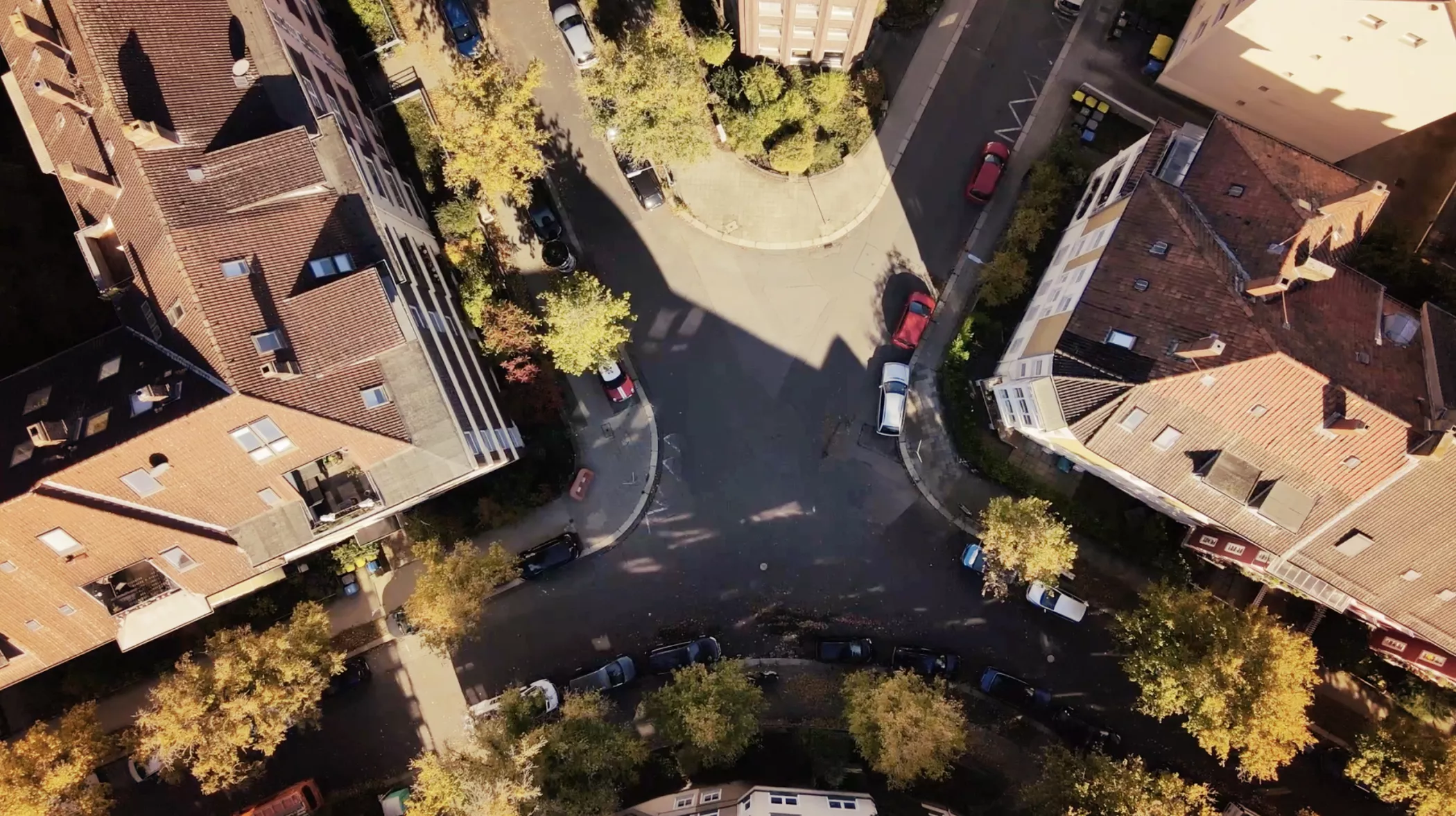 Eine Luftaufnahme einer städtischen Kreuzung, umgeben von Gebäuden, geparkten Autos und Bäumen, die lange Schatten werfen.