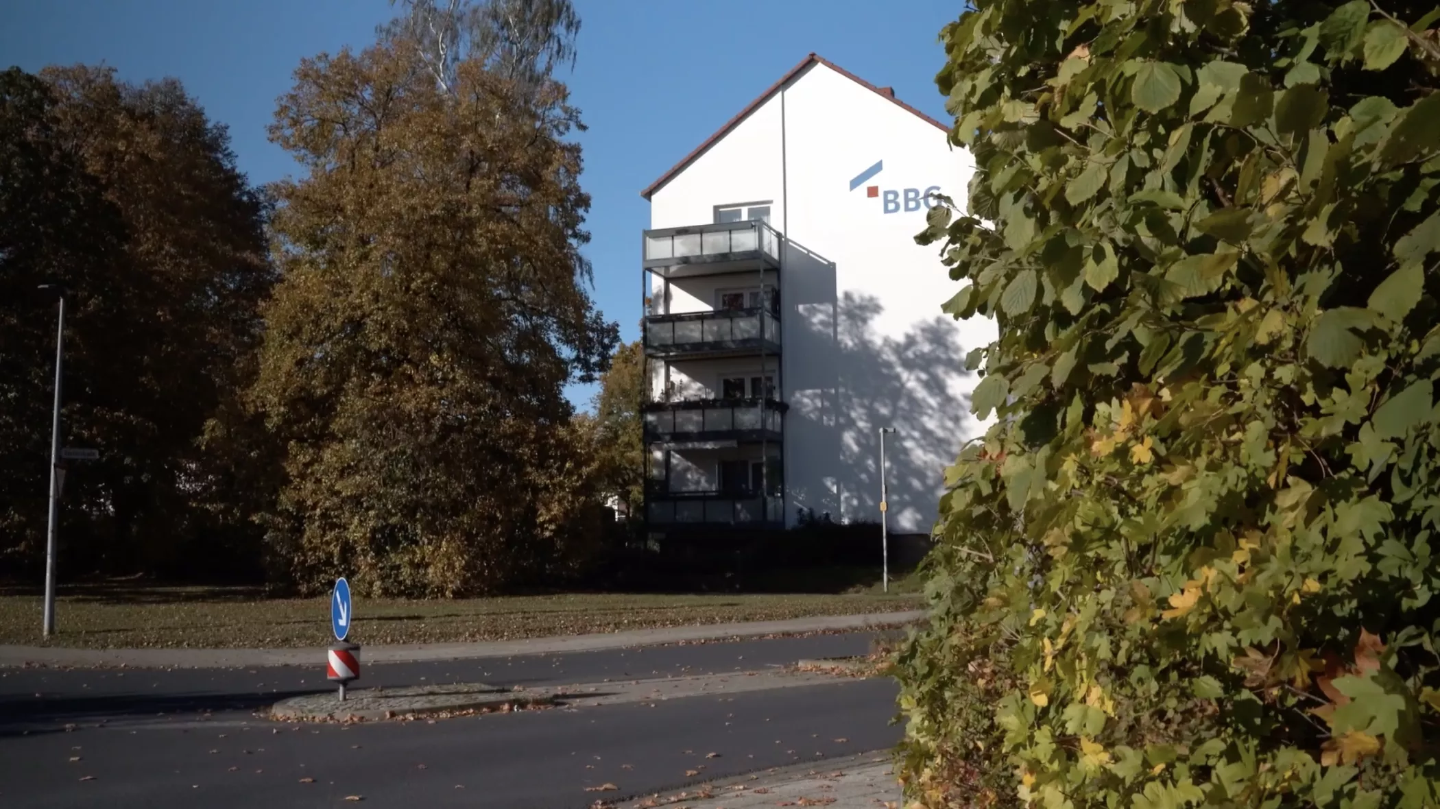 Ein weißes Gebäude mit Balkonen steht an einem sonnigen Tag hinter Bäumen und Büschen in der Nähe einer Straße.