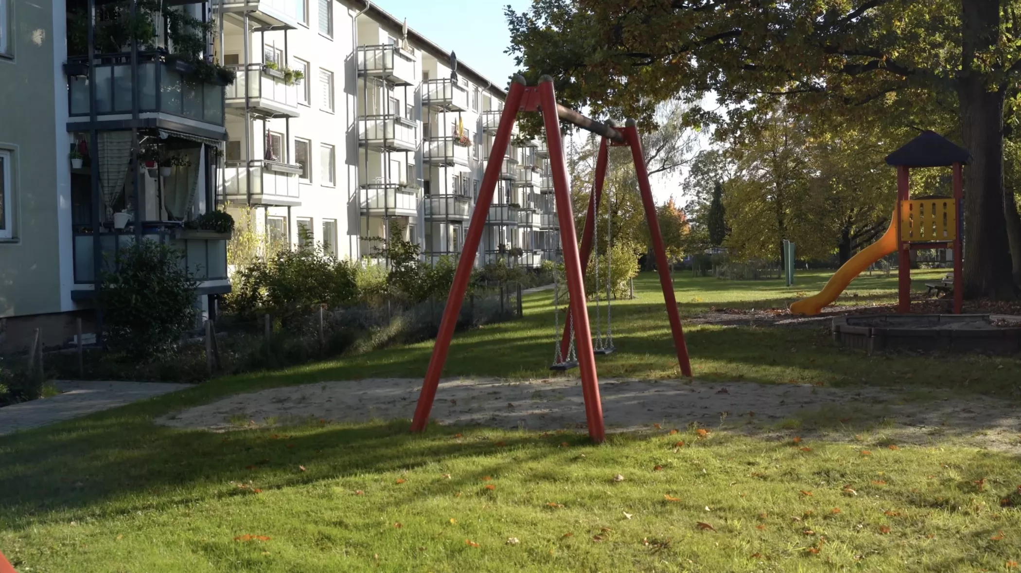 Une balançoire rouge se dresse dans un parc herbeux près d'un immeuble ; on aperçoit des arbres et un toboggan à l'arrière-plan.