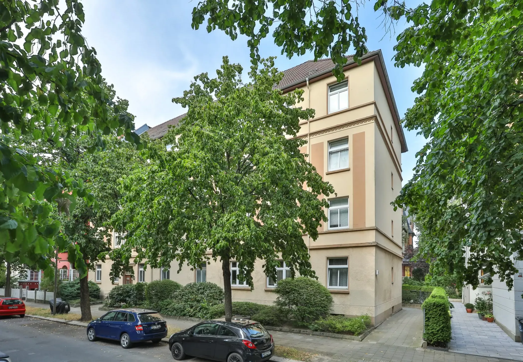 Ein beigefarbenes Wohnhaus mit hohen Fenstern, umgeben von Bäumen und parkenden Autos an einer ruhigen Straße.