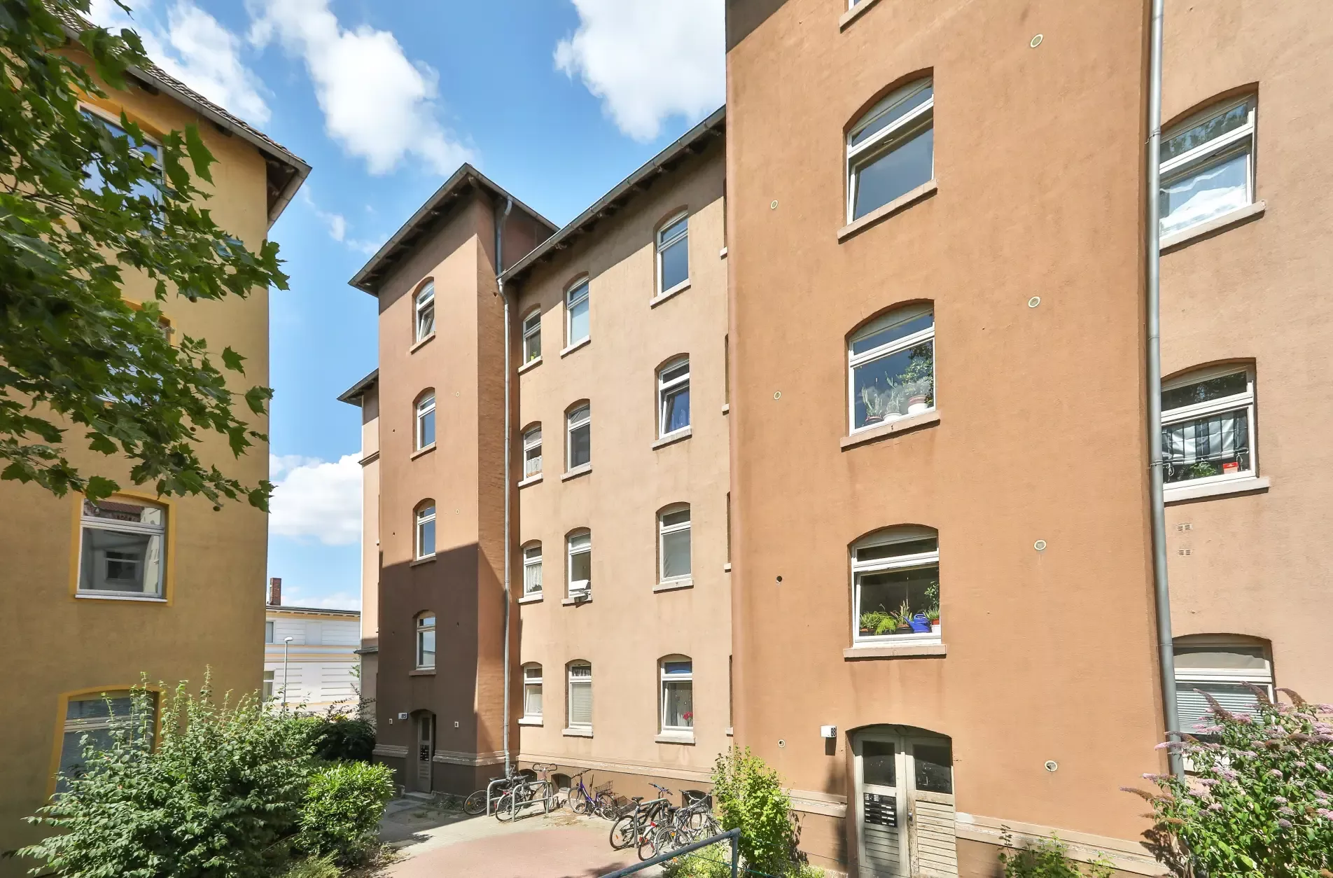 Чотириповерховий коричневий житловий будинок з багатьма вікнами, деякі з них засаджені рослинами, а біля входу в сонячний день припарковані велосипеди.