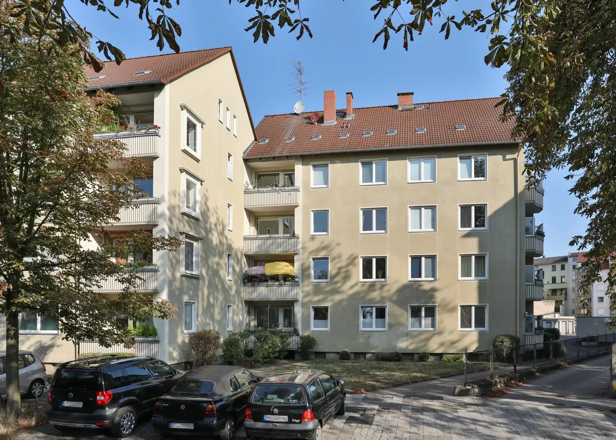 Ein dreistöckiges beigefarbenes Wohnhaus mit Balkonen, davor geparkten Autos und Bäumen, die an einem sonnigen Tag Schatten werfen.