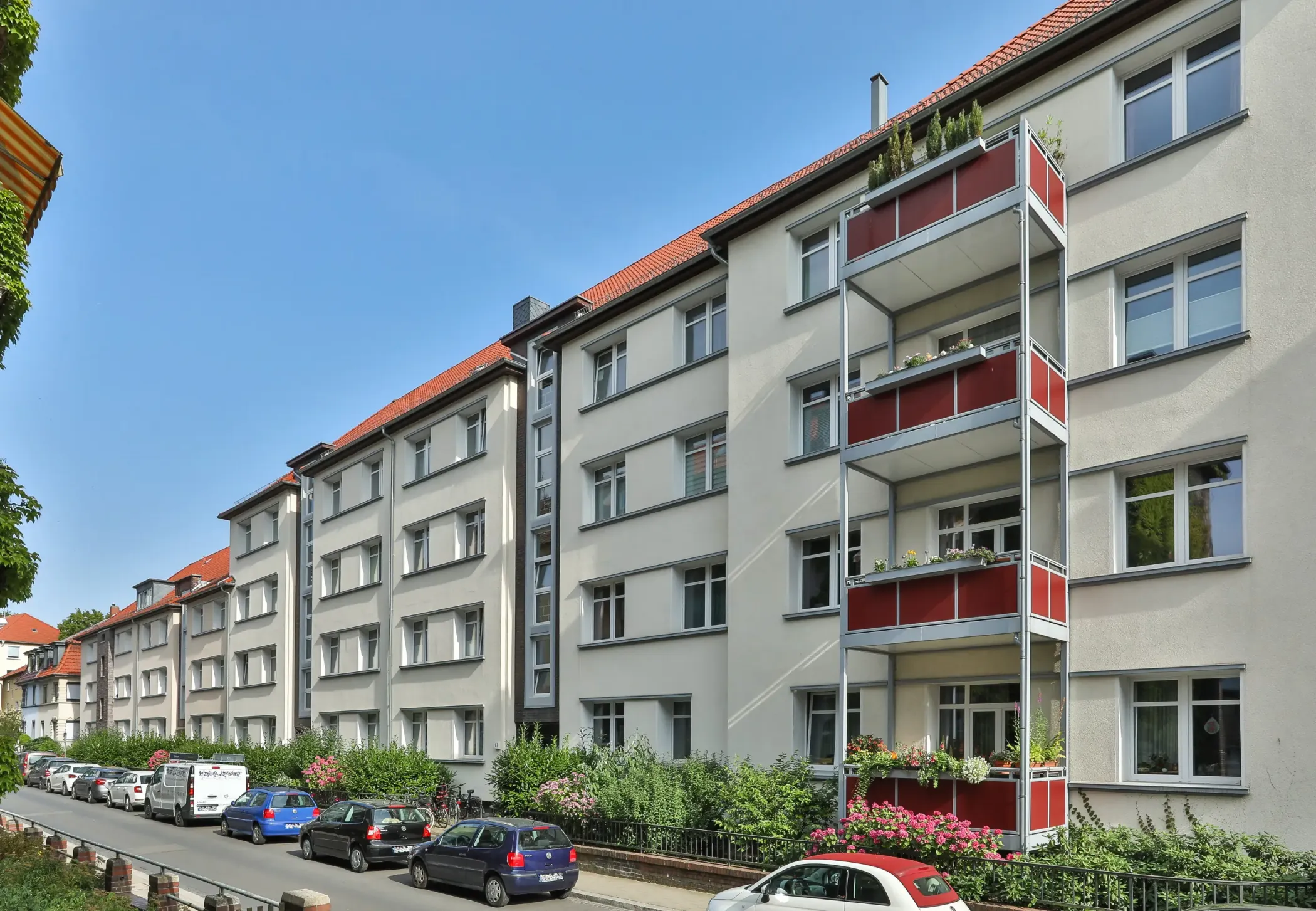 Un immeuble de quatre étages avec des balcons, des accents rouges et des voitures garées le long de la rue, sous un ciel bleu clair.