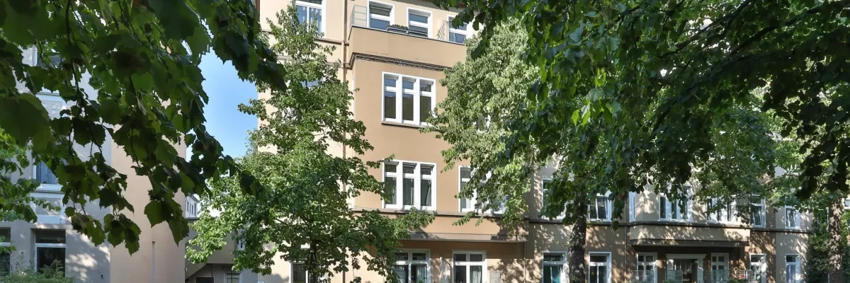 Ein beigefarbenes Wohnhaus mit weiß verzierten Fenstern, Bäumen davor und parkenden Autos entlang der Straße an einem sonnigen Tag.