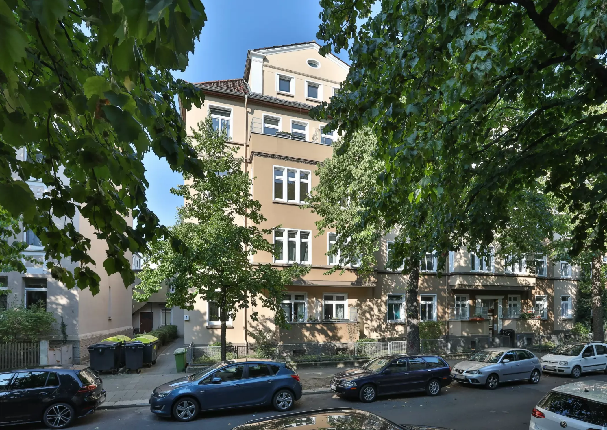 Ein beigefarbenes Wohnhaus mit weiß verzierten Fenstern, Bäumen davor und parkenden Autos entlang der Straße an einem sonnigen Tag.
