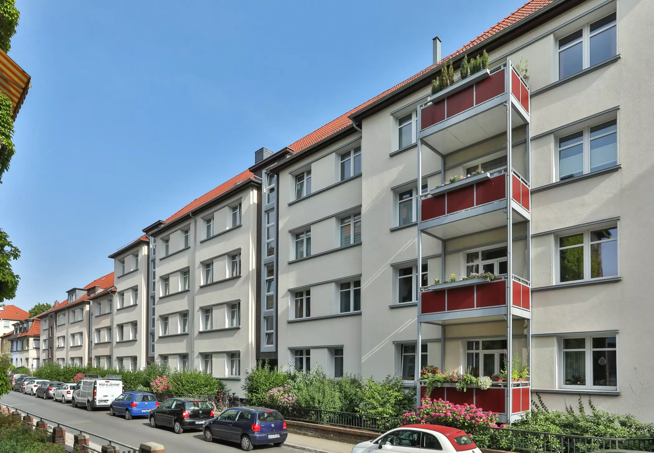 Ein vierstöckiges Wohnhaus mit Balkonen, roten Akzenten und parkenden Autos, die die Straße davor säumen, unter einem klaren blauen Himmel.