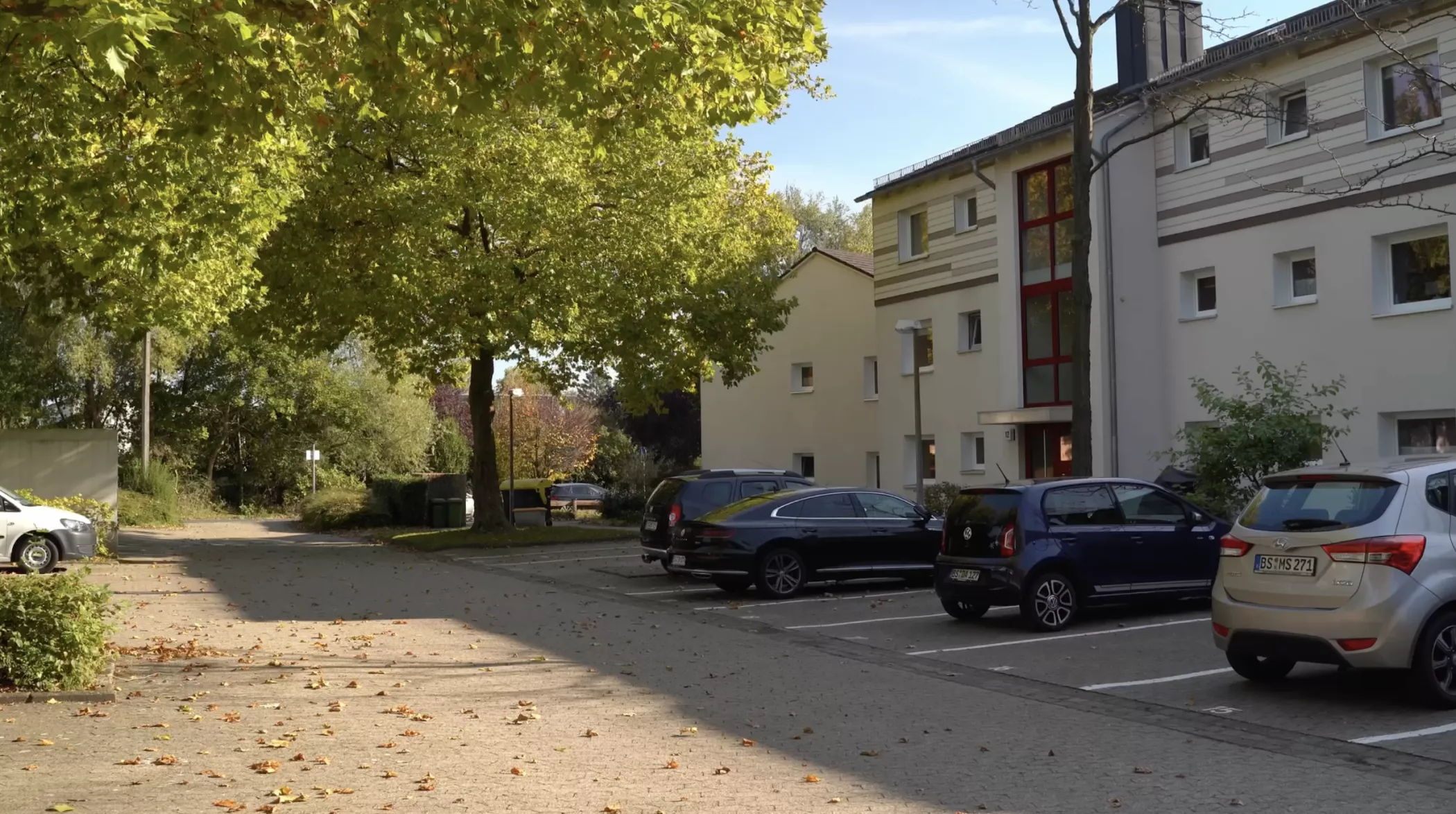 A quiet residential parking lot with several cars, shady trees and residential buildings under a clear sky.