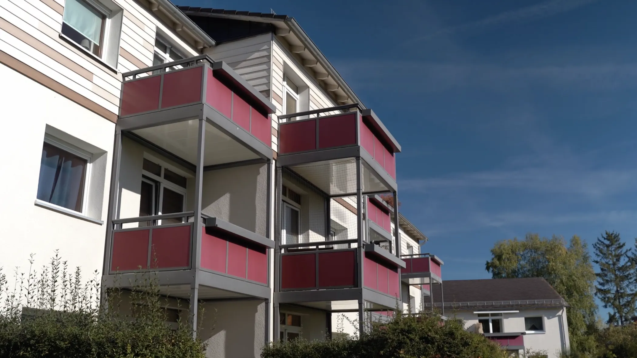 Ein dreistöckiges Wohnhaus mit rot gefärbten Balkongeländern, weißer Verkleidung und großen Fenstern unter einem klaren blauen Himmel.