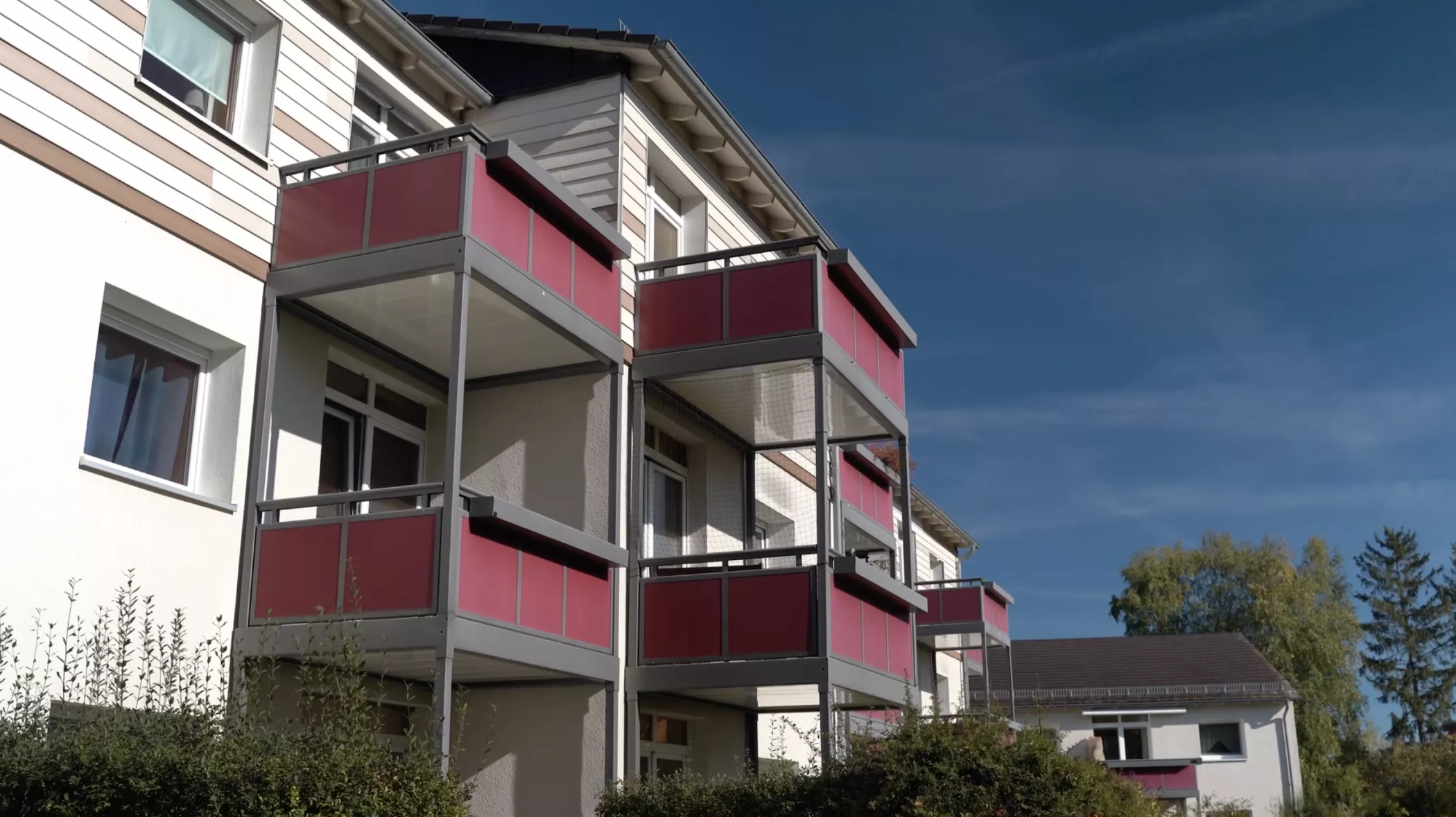 A three-storey residential building with red-colored balcony railings, white cladding and large windows under a clear blue sky.