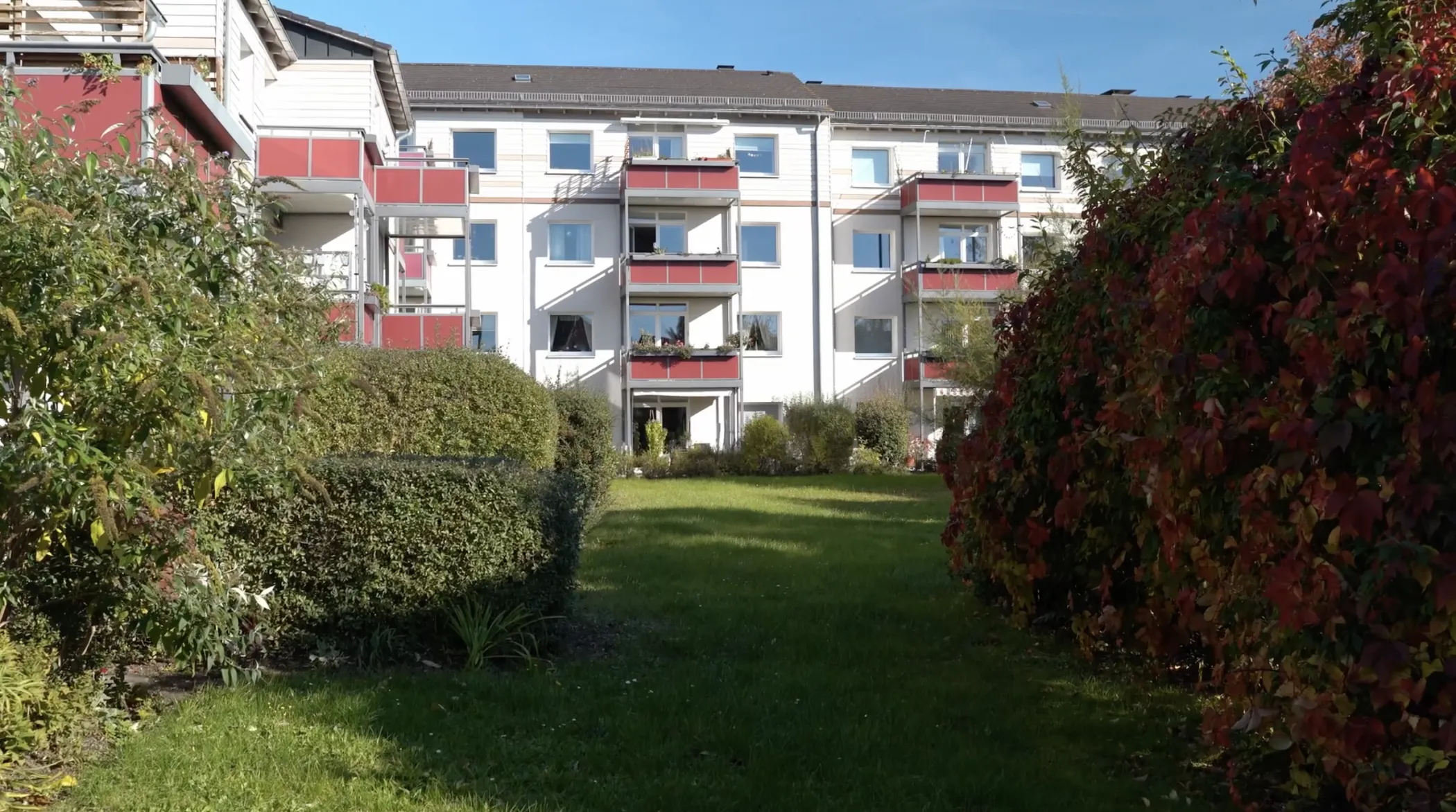 Ein Wohnhaus mit weißen Wänden und roten Balkonen, umgeben von grünen Büschen und Gras unter einem klaren blauen Himmel.