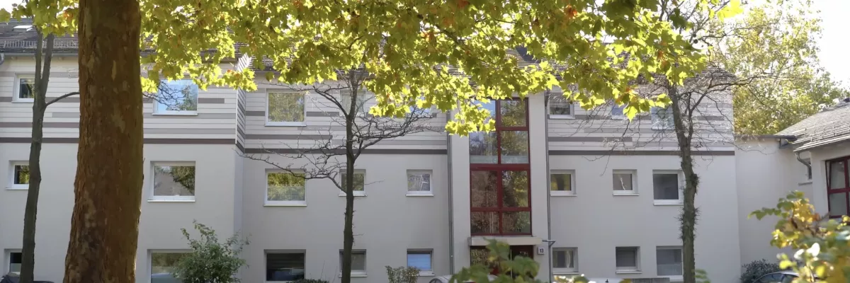 Ein modernes beigefarbenes Wohnhaus mit großen Fenstern, das durch die üppig grünen Bäume und die davor geparkten Autos sichtbar ist.