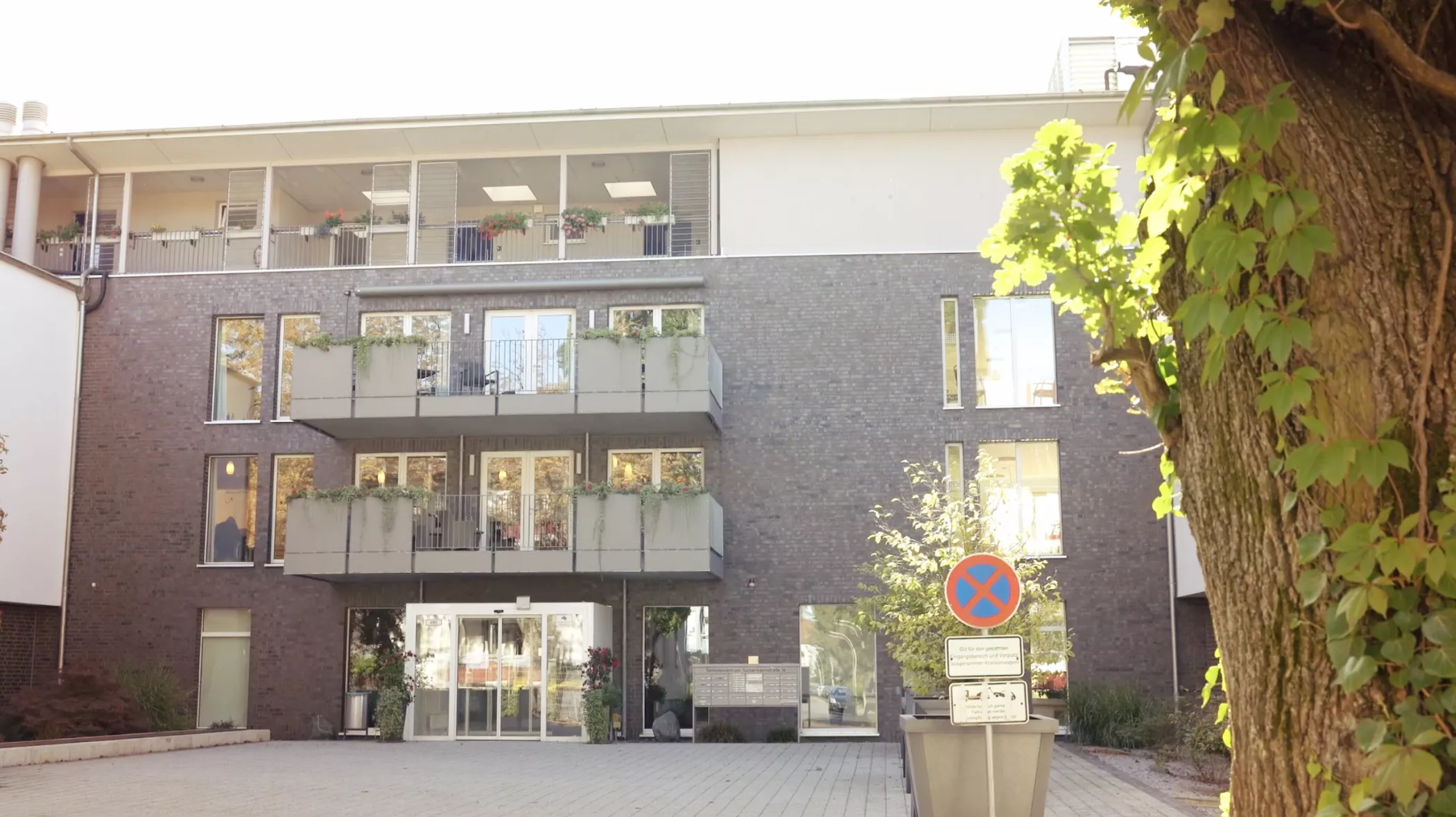 Ein dreistöckiges modernes Backsteingebäude mit Balkonen, großen Fenstern und einem Parkverbotsschild davor, umgeben von Bäumen.