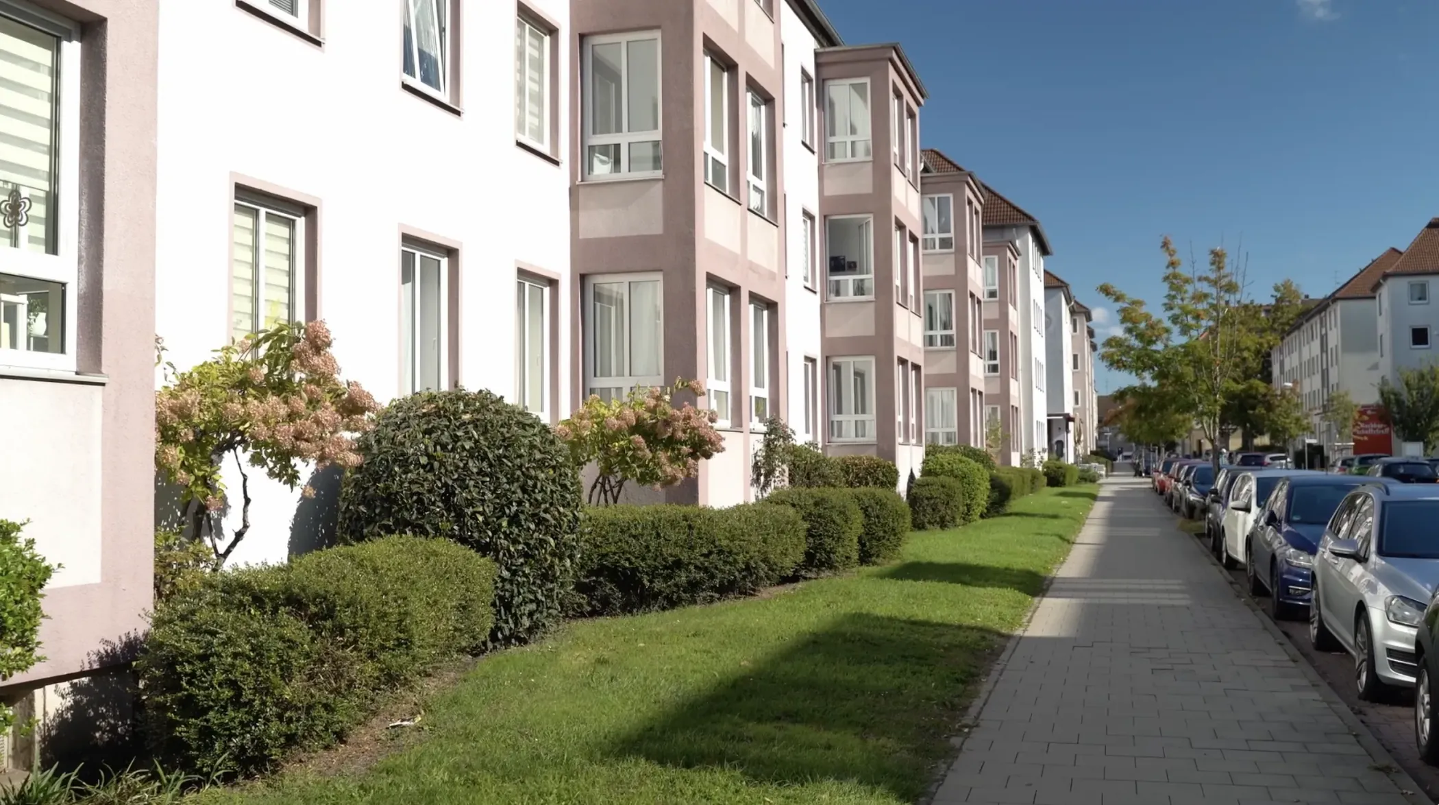 Une rangée d'immeubles modernes avec de grandes fenêtres, des arbustes et un trottoir sous un ciel bleu clair.