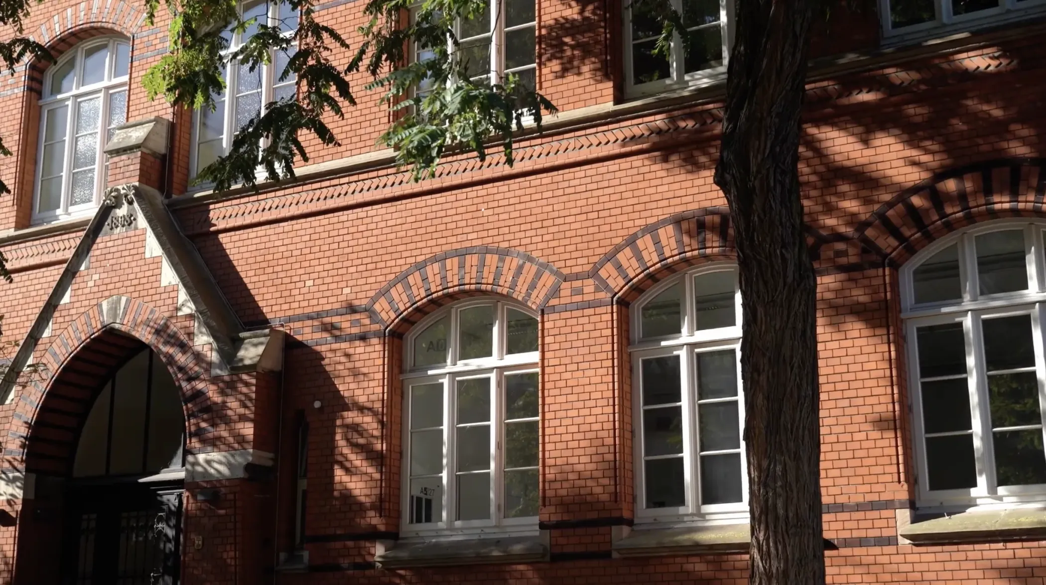 Un bâtiment en briques rouges avec des fenêtres cintrées et un arbre devant lui, le soleil projetant des ombres sur la façade.