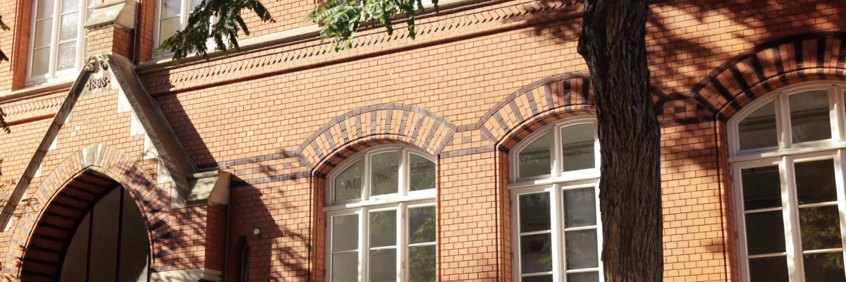 Ein rotes Backsteingebäude mit großen, weiß gerahmten Fenstern, gewölbten Backsteindetails und einem Baum, der die Fassade teilweise beschattet.