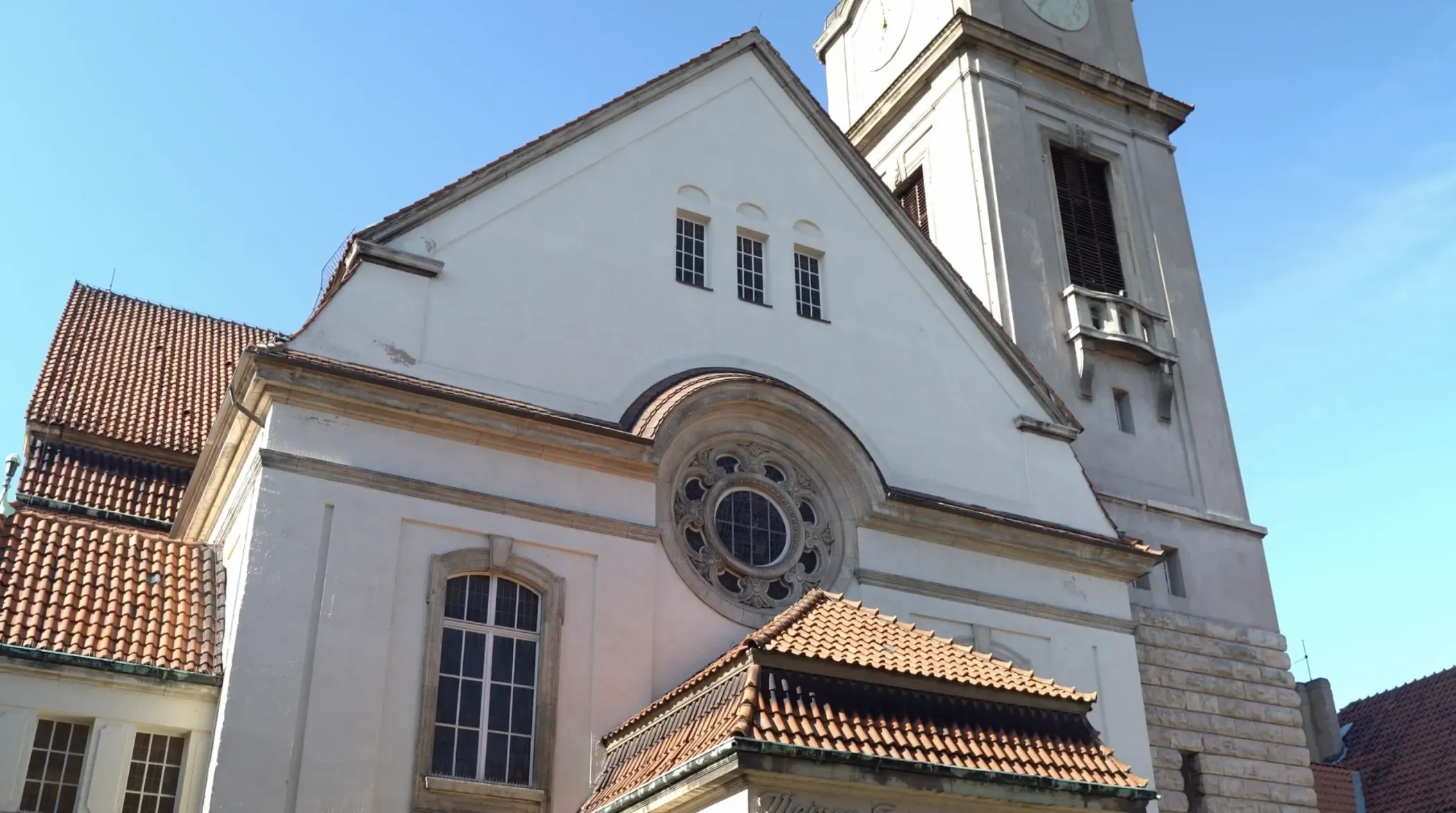 Une église blanche avec un clocher, des fenêtres cintrées et des toits de tuiles rouges dans un ciel bleu clair.