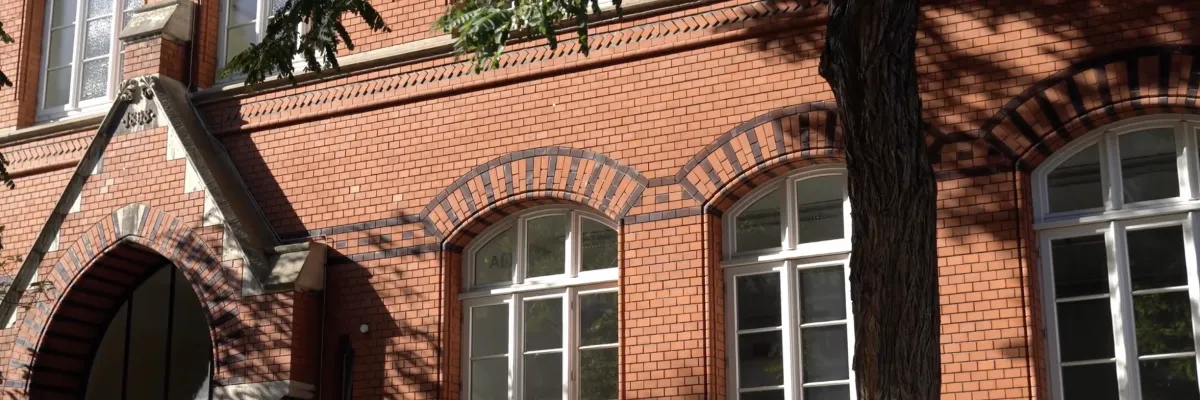 A red brick building with arched windows in a white frame, partially shaded by a large tree on the façade.