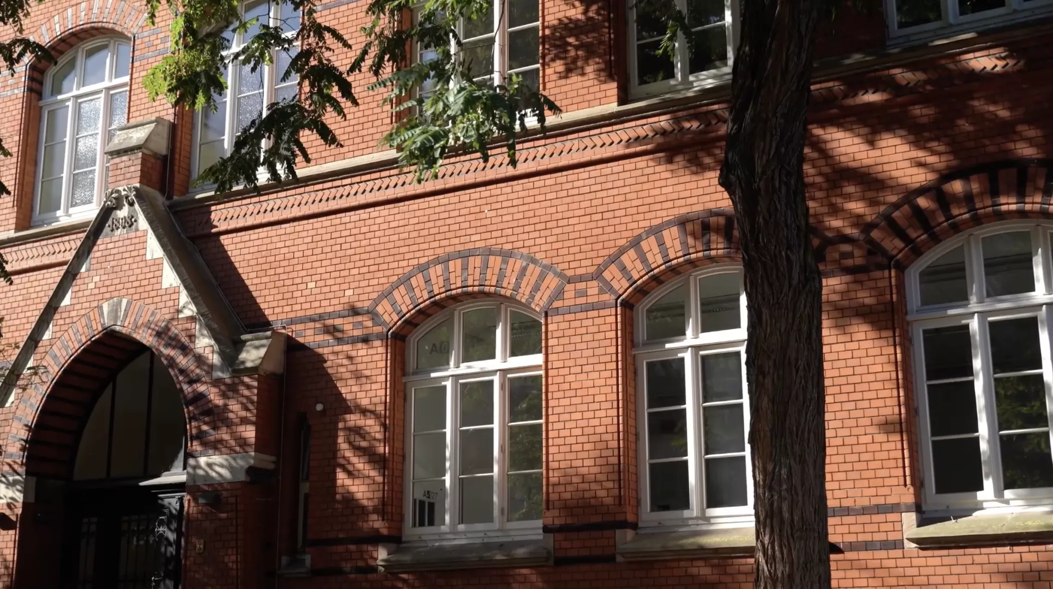 Kemerli pencereleri olan kırmızı tuğlalı bir bina ve önünde bir ağaç, güneş ön cepheye gölgeler düşürüyor.