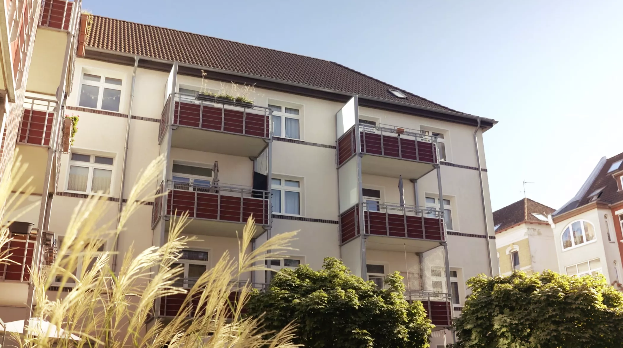 Ein dreistöckiges Wohnhaus mit Balkonen und roten Balustraden, umgeben von Grün unter einem klaren blauen Himmel.