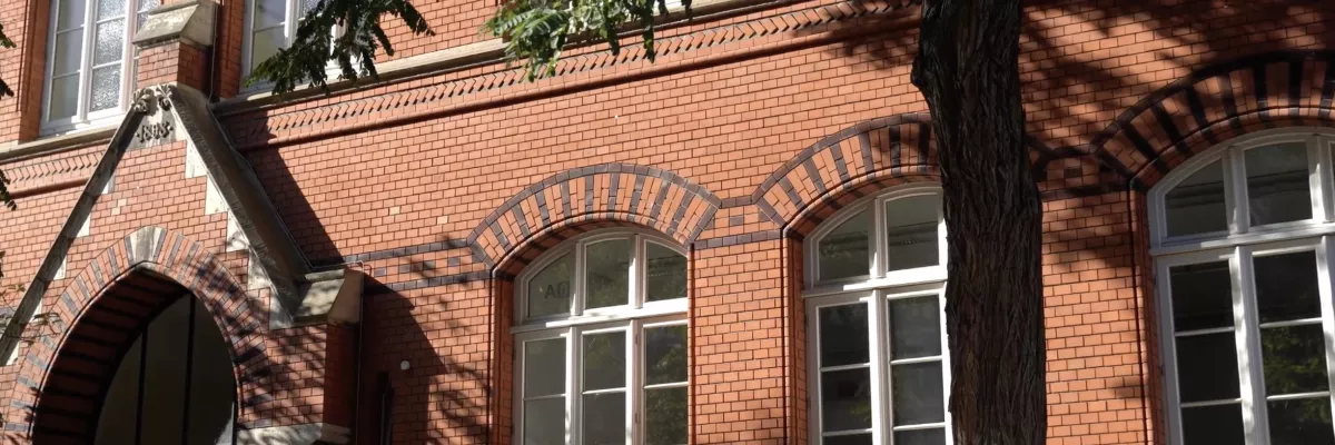 Beyaz çerçeve içinde kemerli pencereleri olan kırmızı tuğlalı bina, ön cephesinde büyük bir ağaç tarafından kısmen gölgelenmiştir.