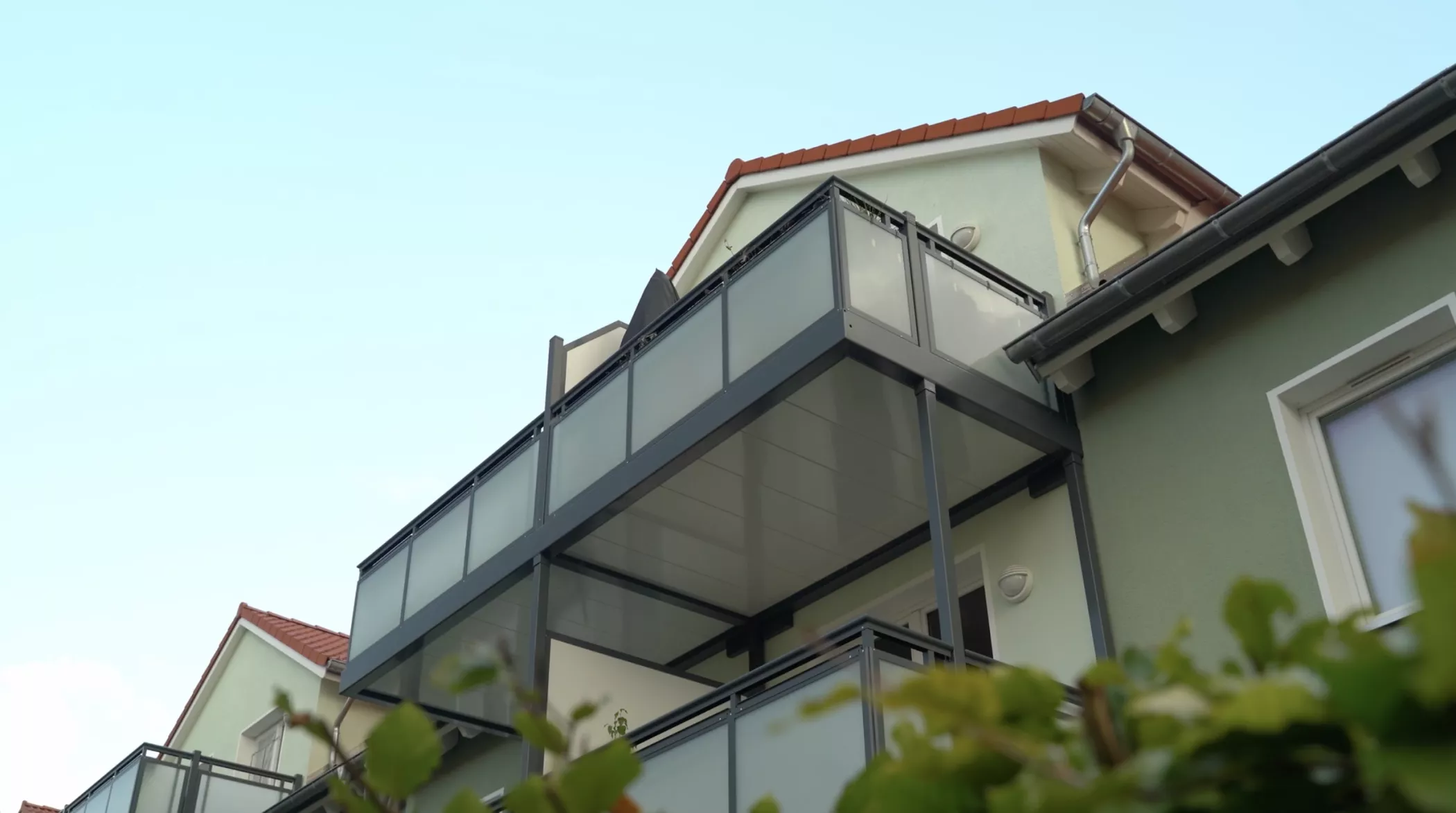 Low angle view of a modern house with glass balconies, green wall and red tiled roof, surrounded by green leaves in the foreground.