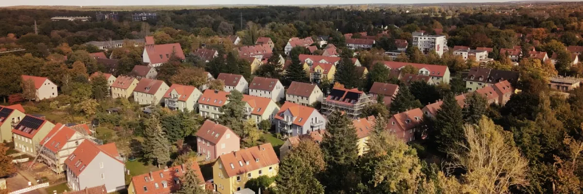 Luftaufnahme eines Vorstadtviertels mit rotbedachten Häusern, umgeben von Bäumen und Grünanlagen unter einem teilweise bewölkten Himmel.