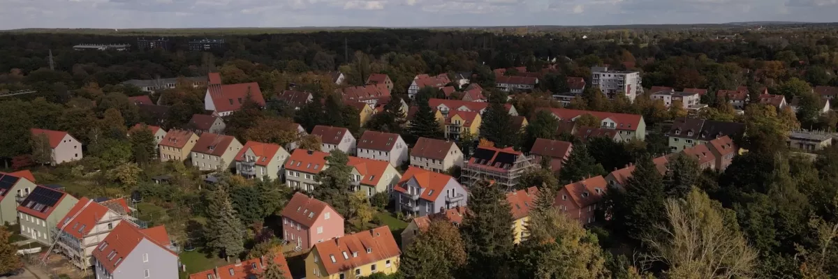 Parçalı bulutlu bir gökyüzü altında ağaçlarla çevrili kırmızı çatılı evlerin bulunduğu bir banliyö bölgesinin havadan görünümü.