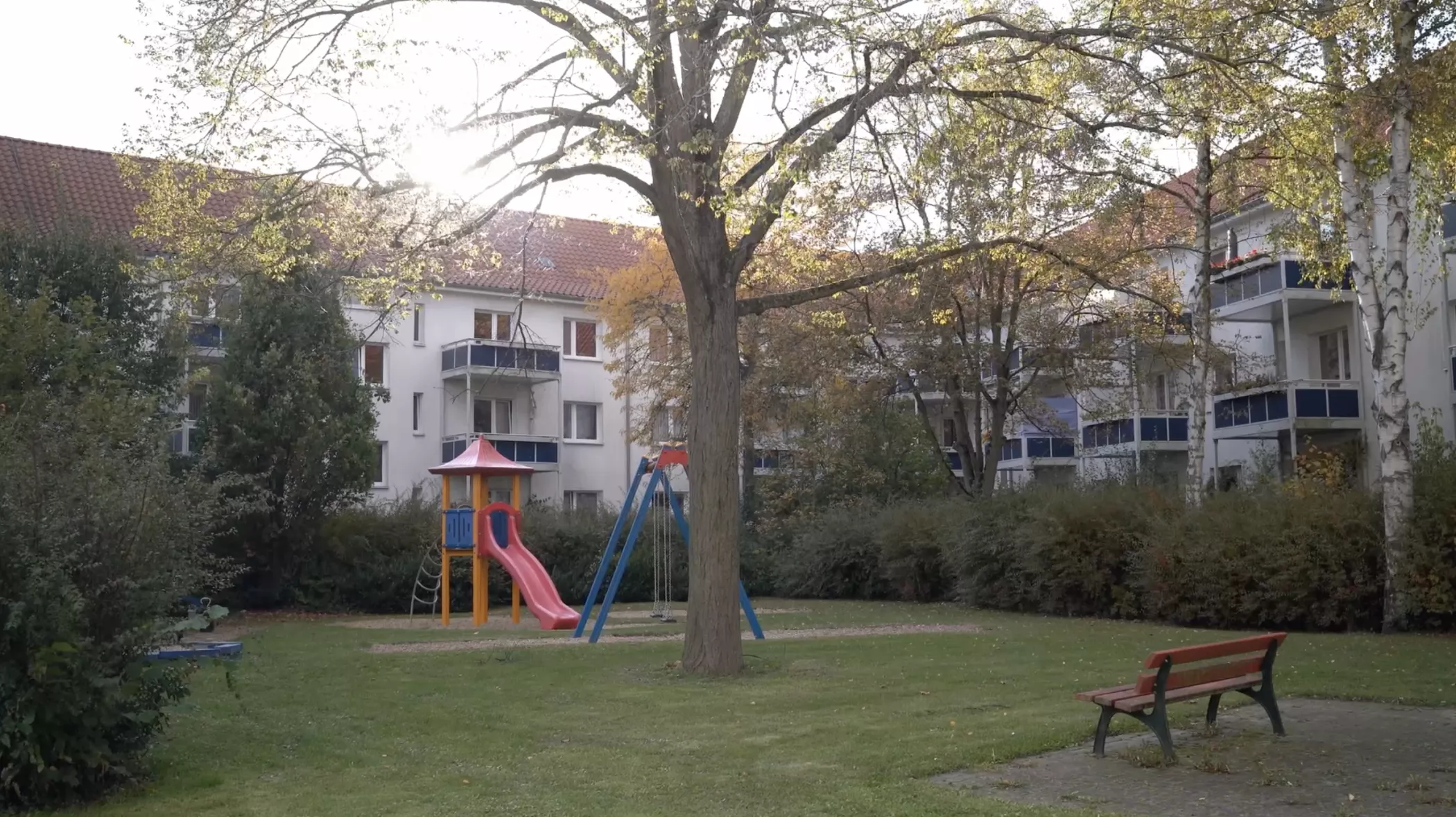 Ein Spielplatz mit einer Rutsche und einer Schaukel im Gras in der Nähe eines Baumes, mit Wohnhäusern und einer roten Bank im Hintergrund.