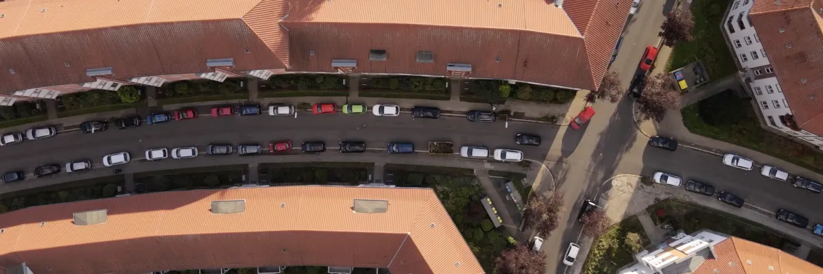 An aerial view of residential buildings with red roofs, a winding road and parked cars in a green neighborhood.